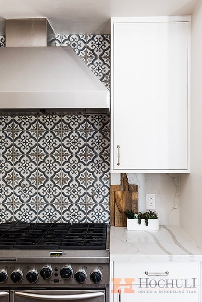 Modern kitchen detail featuring a stainless steel stove, ornate black and white backsplash, and white cabinetry.