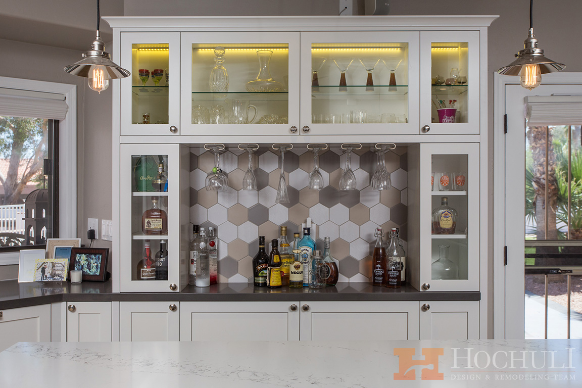 Modern kitchen bar area with glass-fronted cabinets displaying glassware, under-cabinet lighting, and a marble countertop.